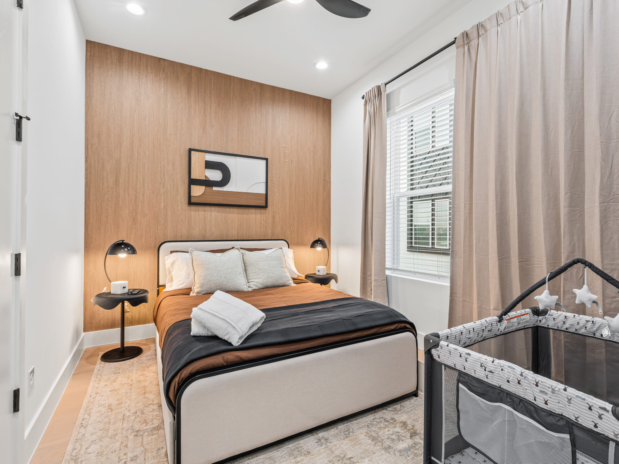 A clean, modern room with a plush queen bed, warm lighting, and a convenient baby playpen for comfortable family stays