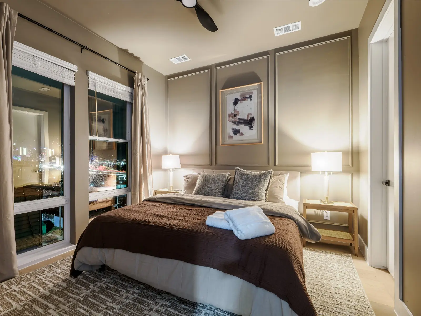 A cozy master bedroom with soft neutral linens, ambient lighting, refined décor, and large windows showcasing a sparkling city view for a hotel-like stay.